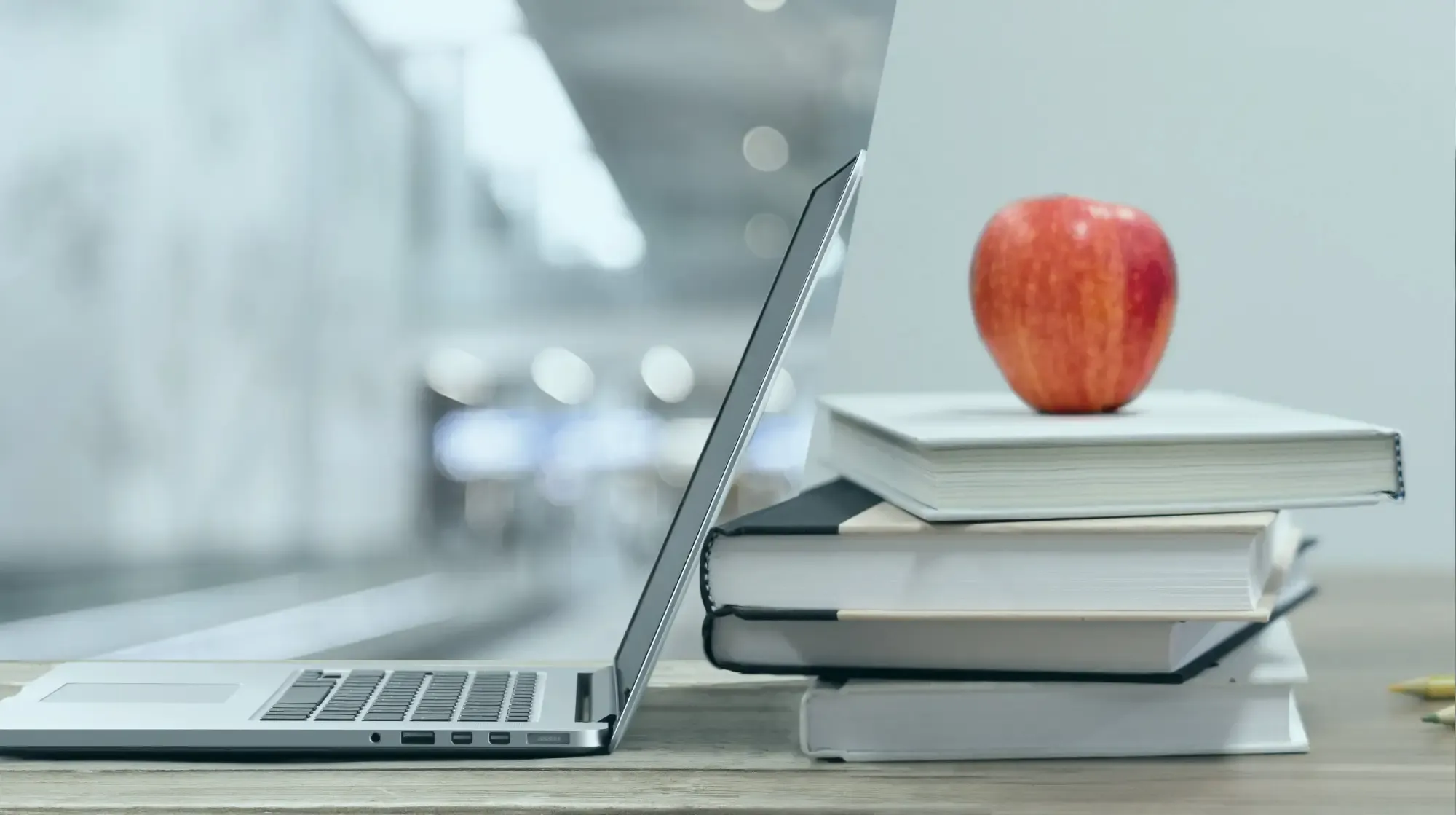 Geöffneter Laptop neben gestapelten Büchern mit einem roten Apfel darauf, auf einem hölzernen Schreibtisch in einer unscharfen Innenumgebung.
