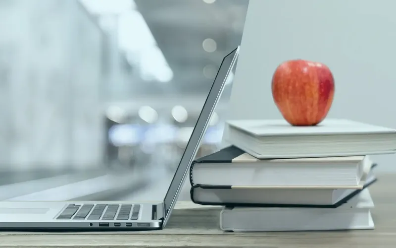 Geöffneter Laptop neben gestapelten Büchern mit einem roten Apfel darauf, auf einem hölzernen Schreibtisch in einer unscharfen Innenumgebung.