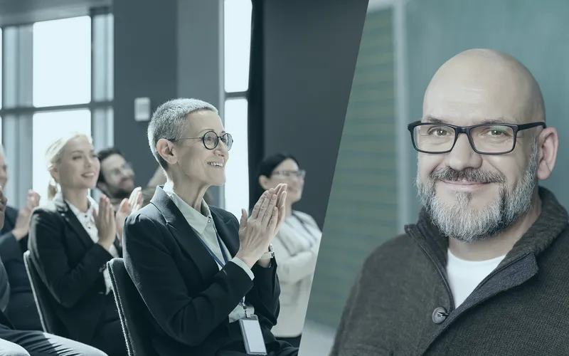 Das Publikum klatscht im Sitzen; rechts steht ein lächelnder Trainer mit Glatze und Brille.